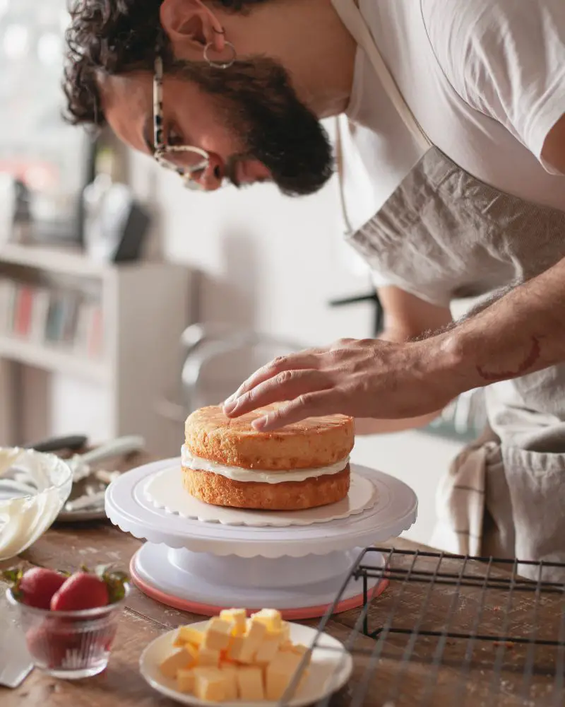 a man in an apron and glasses putting a cake on a cake stand to demonstrate one of many hobbies for men over 60