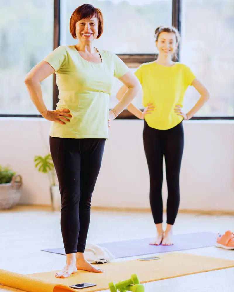 Two women wearing light green and yellow t-shirts and black leggings standing on yoga mats to demonstrate examples of HIIT workouts for women over 50