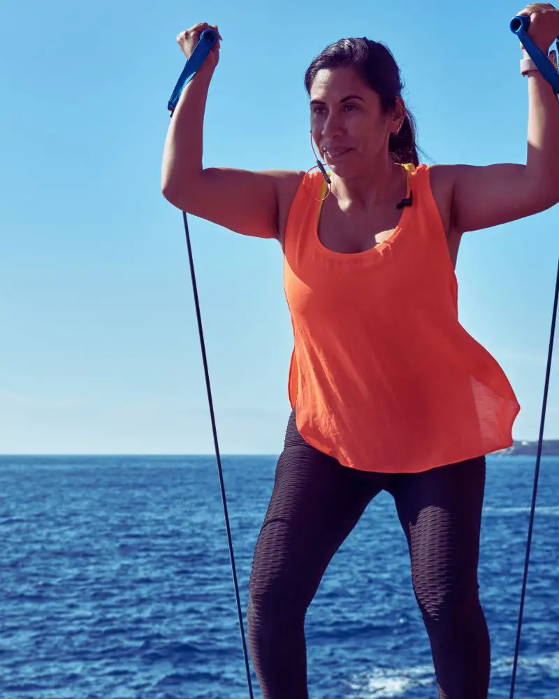 Woman standing by the sea wearing a peach tank top and black leggings while exercising with an exercise band to demonstrate great HIIT workouts for women over 50