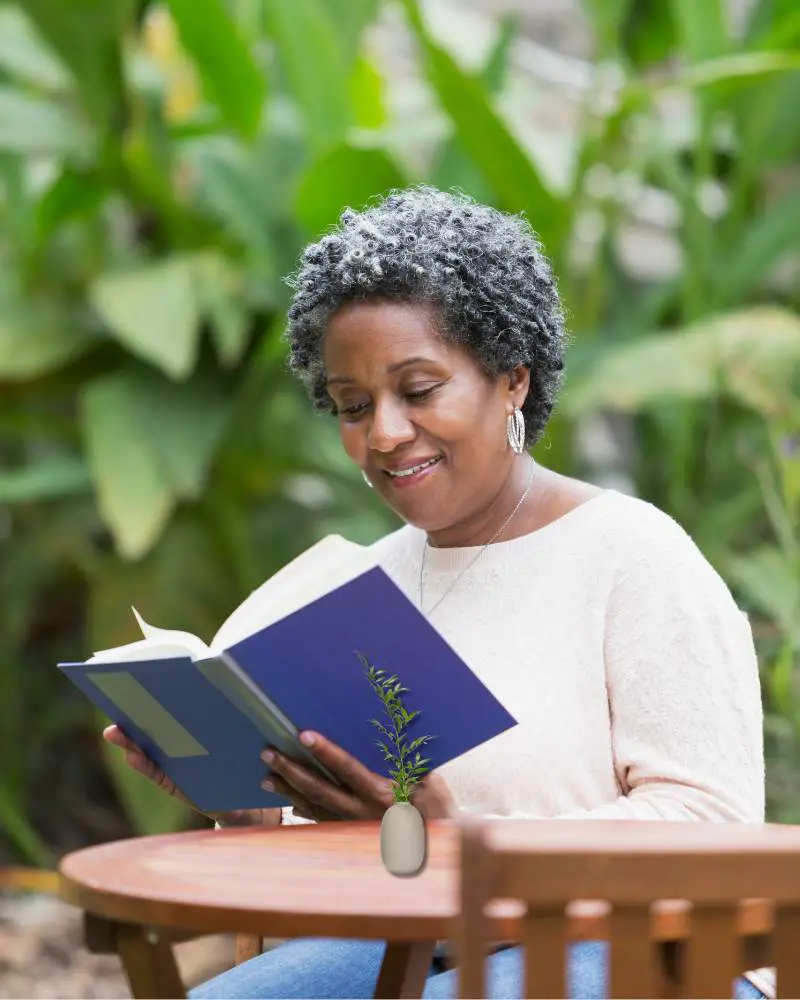 A woman sitting in front of a green tree reading a blue book to demonstrate one of the benefits of Prime for seniors.