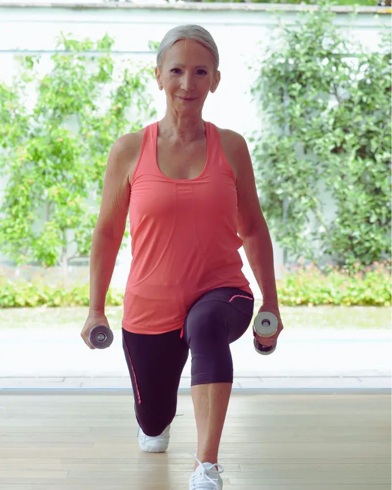 Woman with a peach shirt and black leggings doing a lunge with dumbbells to demonstrate one of many HIIT workouts for women over 50