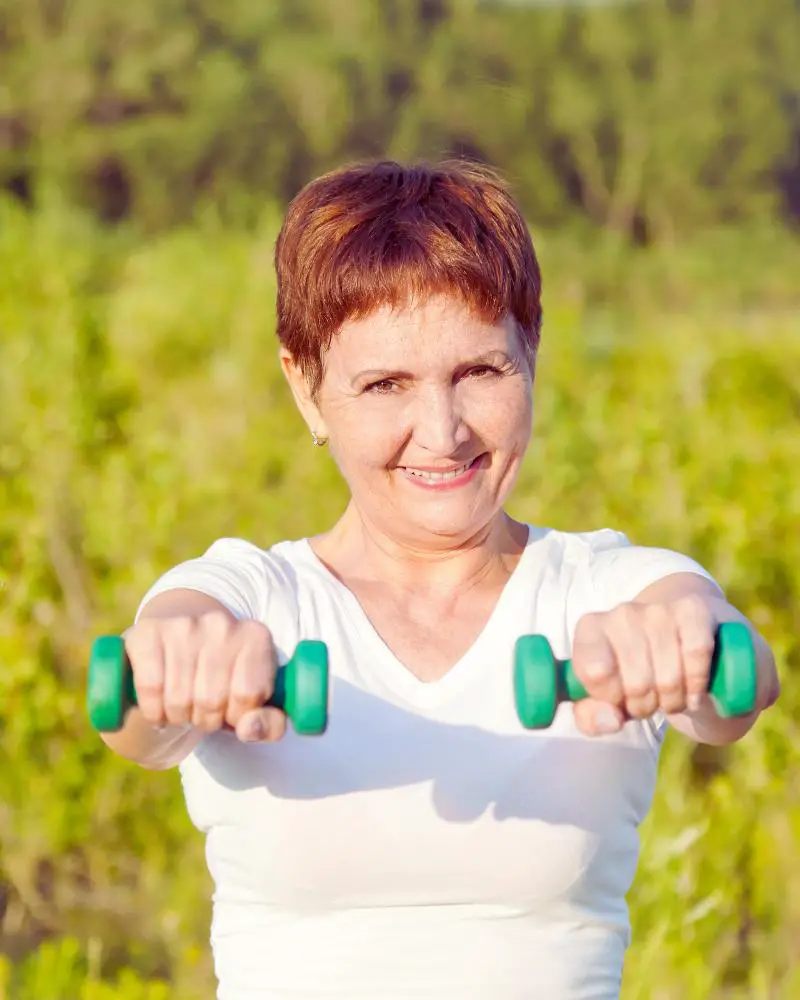 Woman with short red hair standing in a field holding a pair of green dumbbells about to demonstrate a HIIT workout for women over 50.