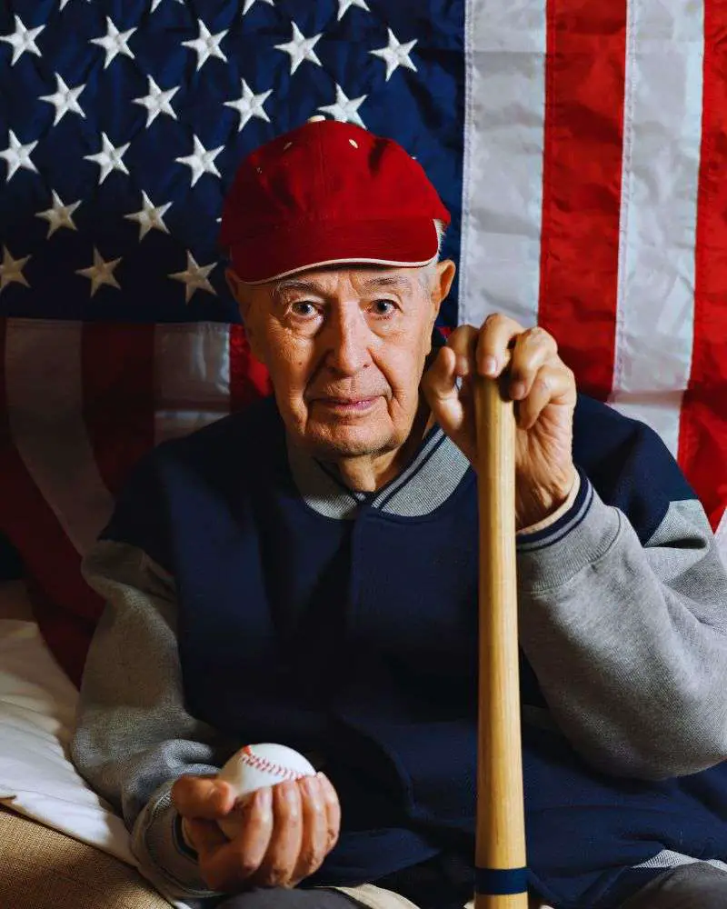 older man in an over 50 baseball league posing in front of the American flag holding a baseball bat.