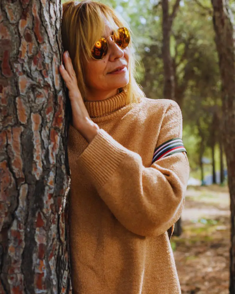 Senior female model wearing a beige sweater leaning against a tree to demonstrate modelling over 50