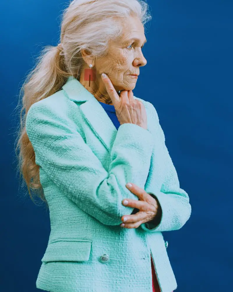 Older woman with long grey hair in a ponytail looking off into the distance, wearing a turquoise blazer to demonstrate over-50-modelling