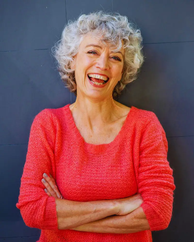 Woman with red sweater leaning against a blue wall with her arms folded. She is smiling after having received an answer to Are Veneers Permanent?