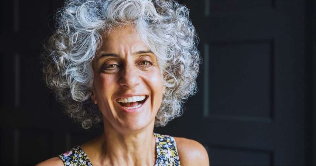 A smiling woman with a head of thick, curly gray hair to demonstrate that she has used the best shampoo and conditioner for over 50 hair.