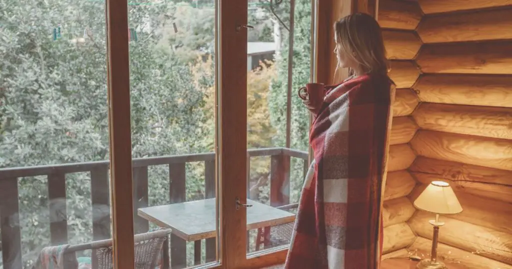 a woman wrapped in a blanket looking out a window to convey the sentiment in cabin quotes for instagram