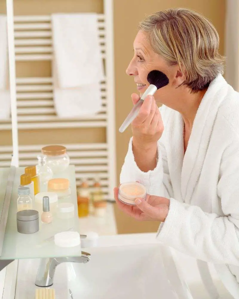 a woman in a bathrobe applying makeup after learning how to conceal under eye bags over 50