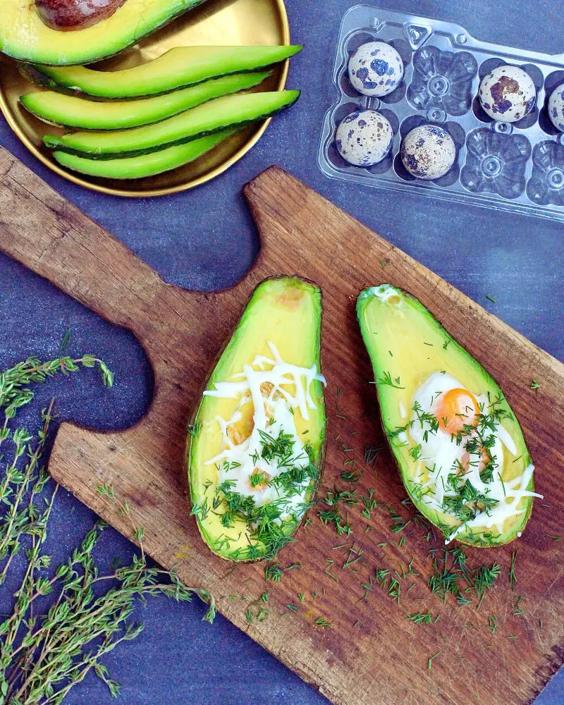 avocado halves with eggs and herbs on a cutting board next to a tray of avocados and a bowl of eggs would complement a post tummy-tuck meal plan